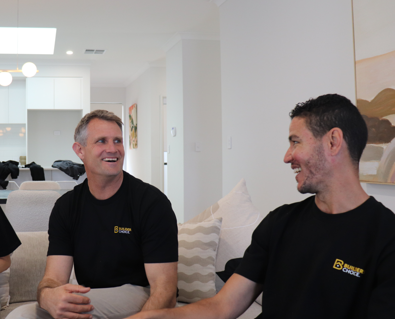 Two Builders Choice team members in branded black t-shirts laughing together on a couch in a display home living room, with an open-plan kitchen visible in the background.