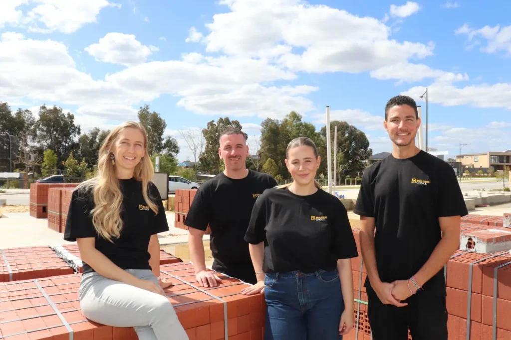 Four Builders Choice team members in branded black t-shirts smiling together outdoors on a construction site, seated and standing on stacked brick pallets with a clear blue sky and trees in the background."